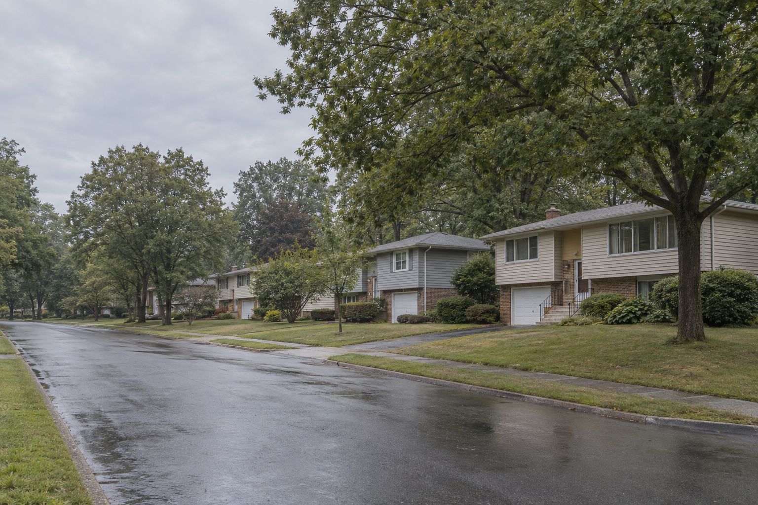 A wet residential street of split-level houses with brick lower floors and single garages, under dense green summer trees on an overcast day