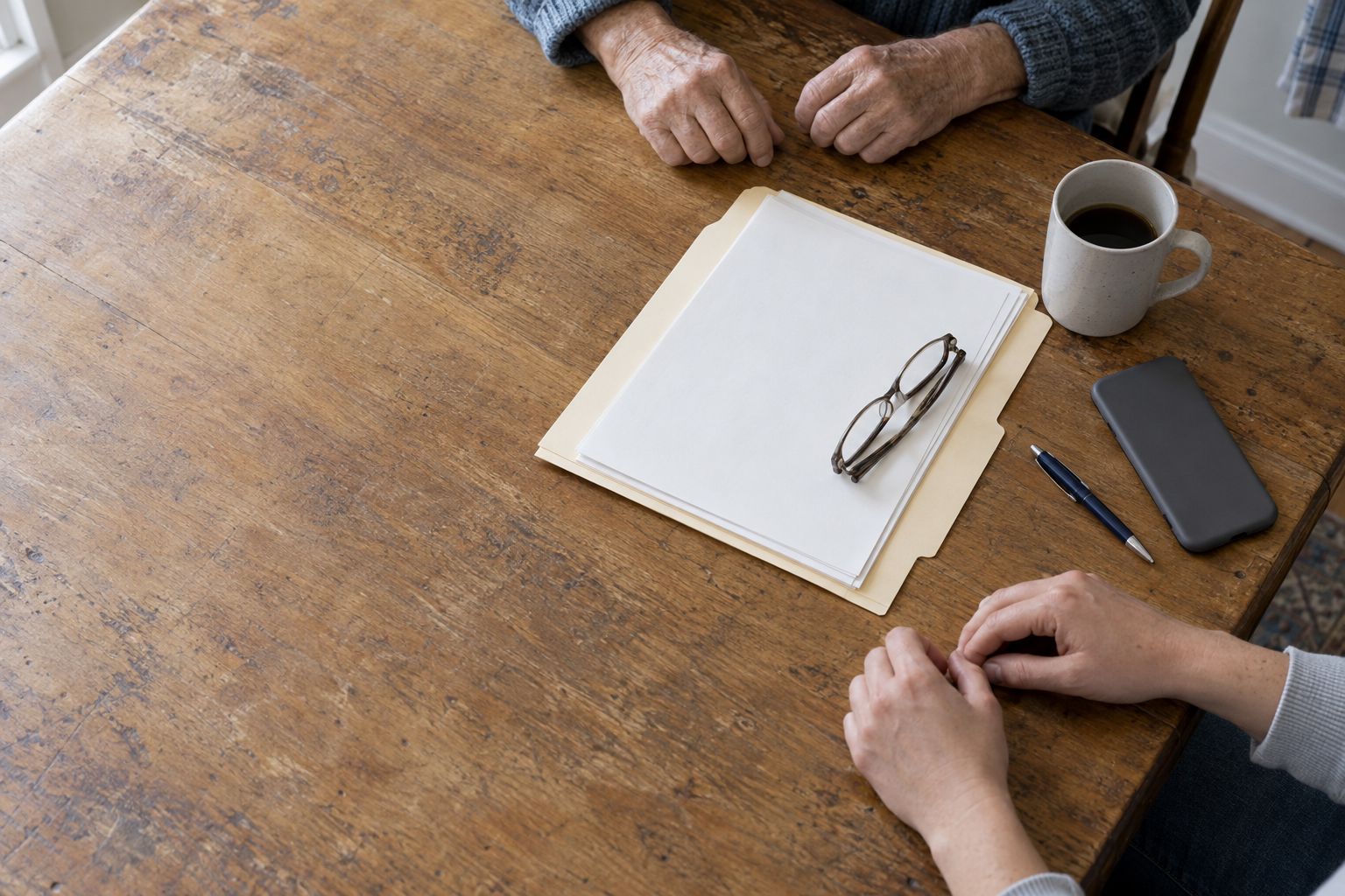 A kitchen table from above with an open folder of papers, reading glasses, a mug and two pairs of hands at the edge