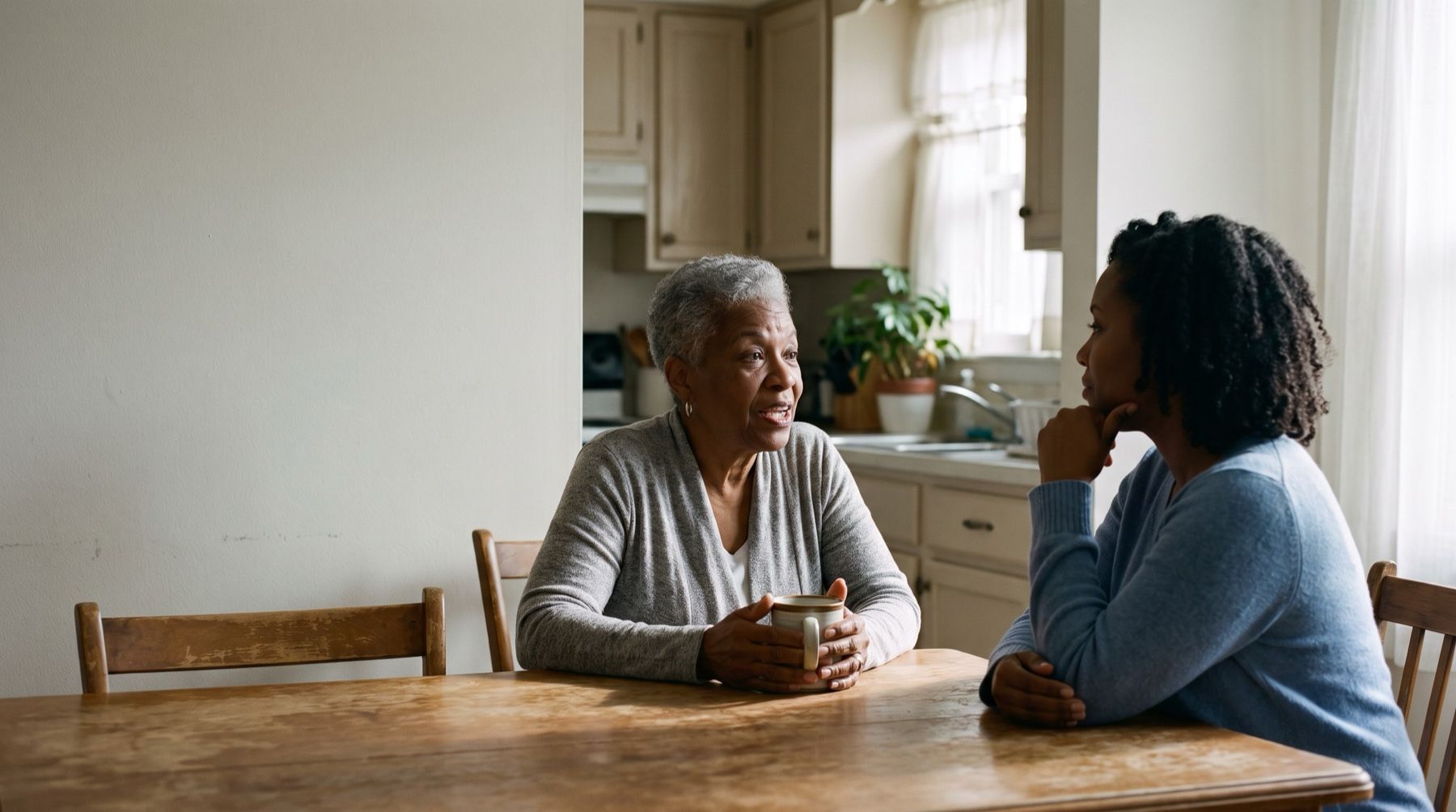An older woman holding a mug and talking with a younger woman across a kitchen table