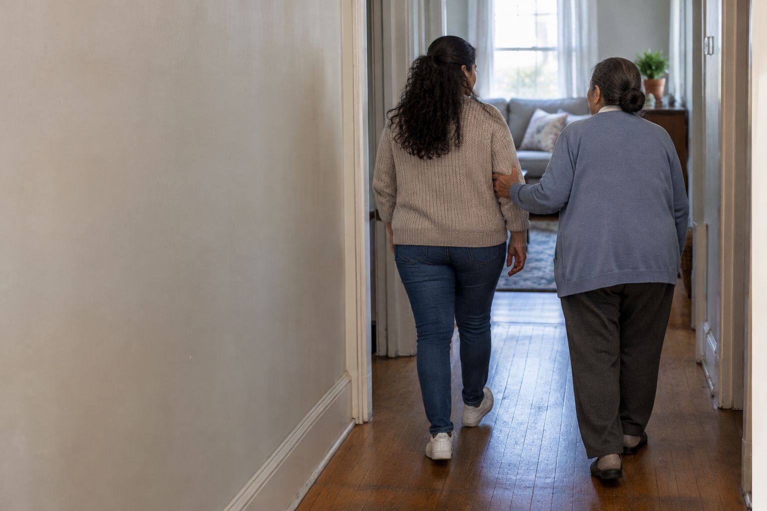 Two women walking slowly along a hallway towards a sunlit room, seen from behind